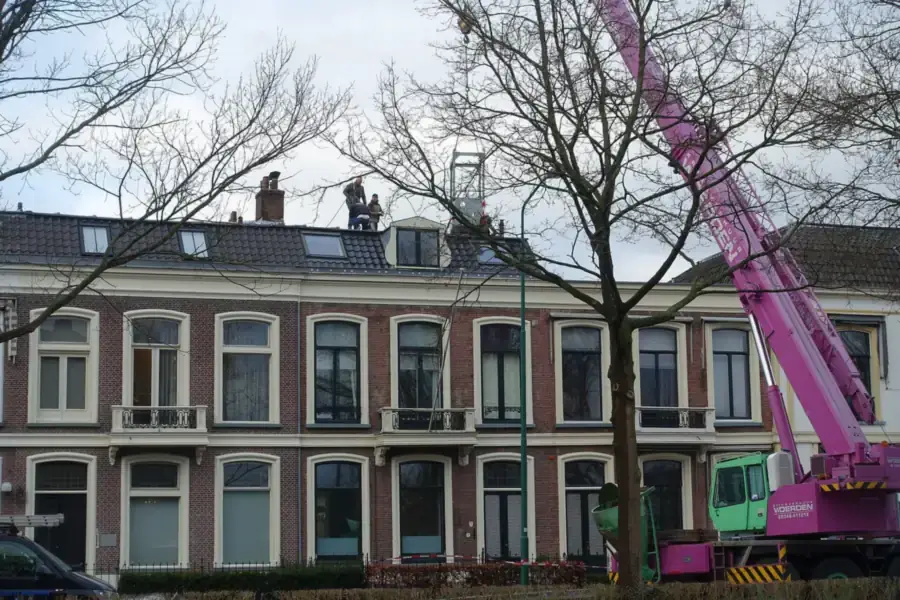 Dakdekkers op het mansardedak van een rij bakstenen herenhuizen met een roze kraan ernaast, herstelwerk na stormschade