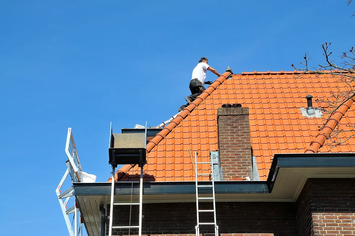Dakdekker zet de nokvorsten vast op een oranje pannendak van een bakstenen woning, herstel van een daklekkage na storm in Amsterdam